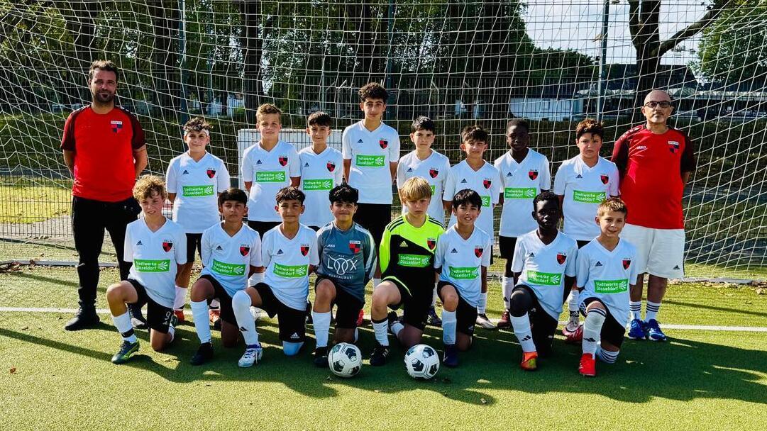 Gruppe von 14 Jugendlichen und zwei Trainern in Fu&szlig;ballkleidung vor einem Tor auf einem Sportplatz.