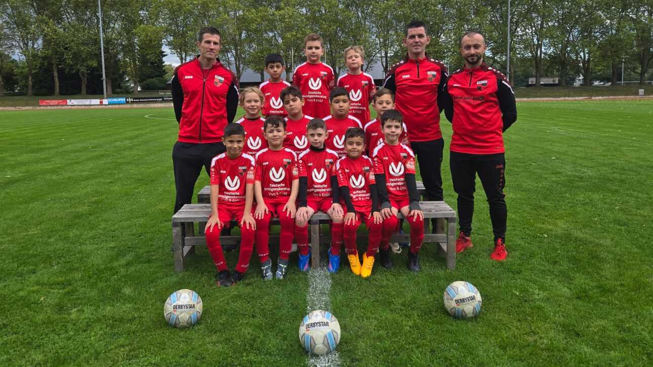 Eine Fu&szlig;ballmannschaft von Kindern in roten Trikots, stehend und sitzend auf einer Bank, mit zwei Trainern auf einem Rasenplatz.