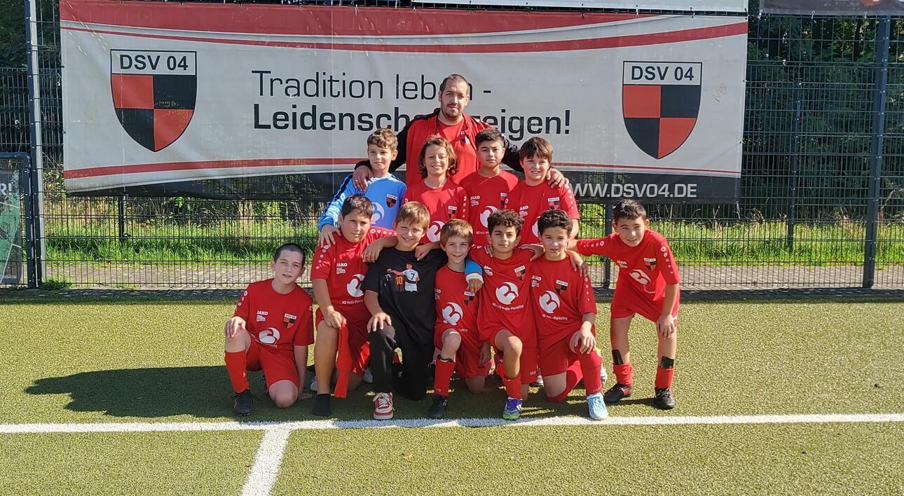 Gruppenfoto einer Fu&szlig;ballmannschaft in roten Trikots, stehend vor einem Banner mit dem Vereinsnamen DSV 04.
