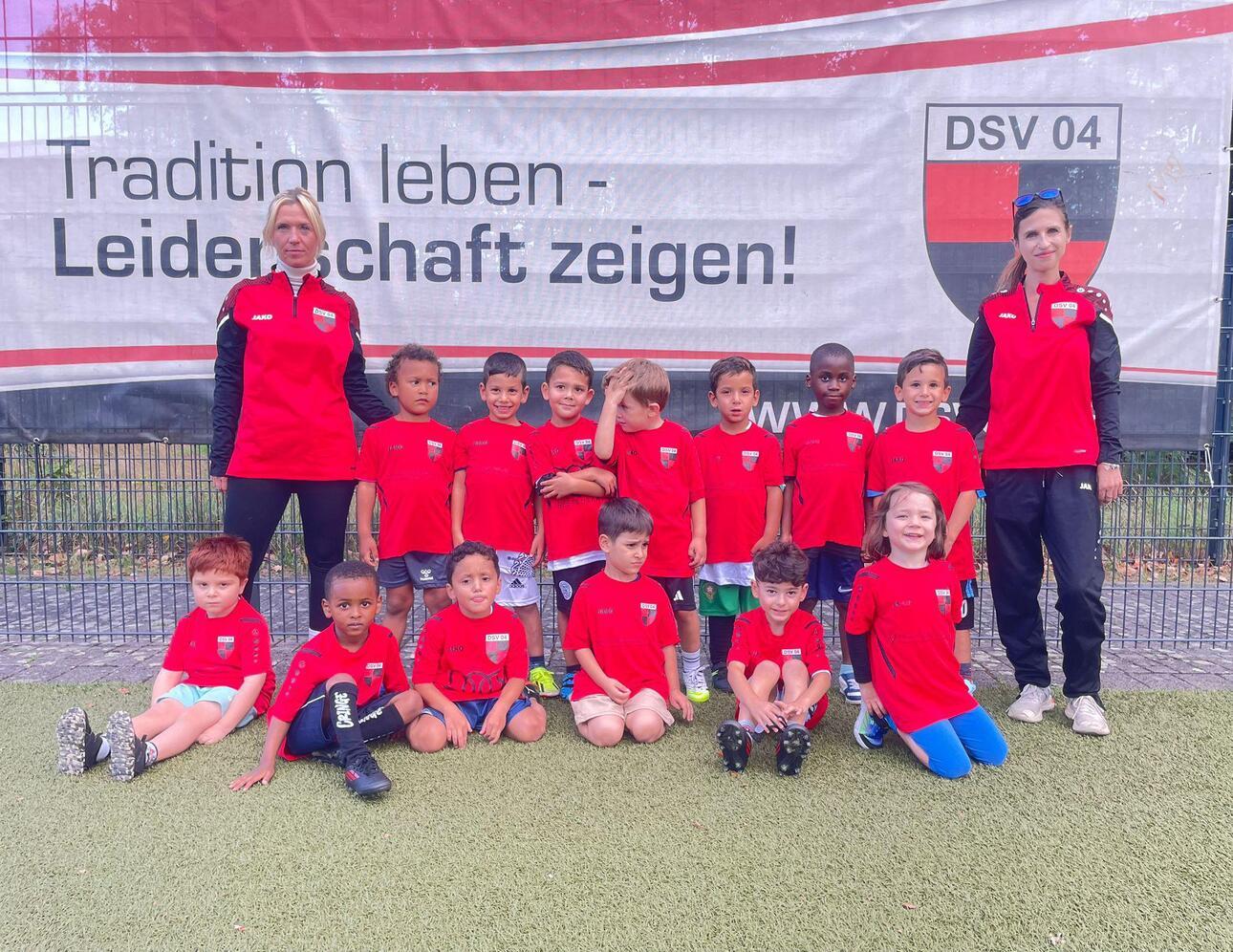 Gruppenfoto von Kindern in roten Trikots, umgeben von zwei Trainerinnen, vor einem DSV 04 Banner auf einem Sportplatz.