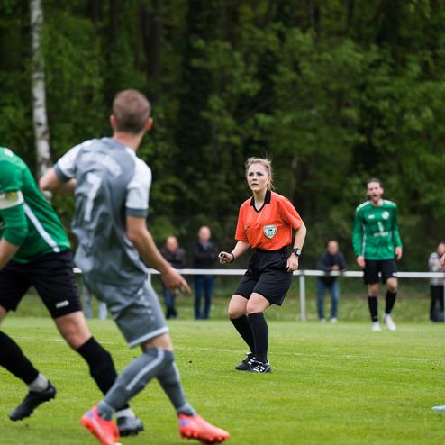 Fu&szlig;ballspiel auf einem Rasenfeld mit Spielern in grauen und gr&uuml;nen Trikots und einer Schiedsrichterin in orange.
