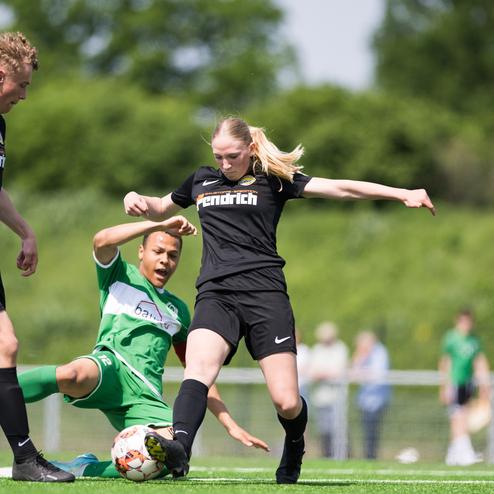Actionreiche Szene im Fu&szlig;ballspiel: Eine Spielerin mit langen Haaren tritt nach einem Ball, w&auml;hrend ein Gegenspieler am Boden liegt.
