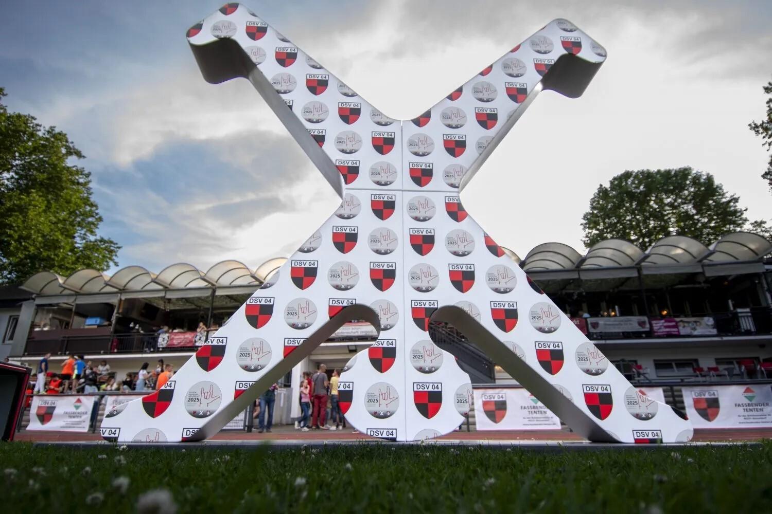 Gro&szlig;es, wei&szlig;es X-Symbol mit Vereinswappen auf einem Sportplatz, umgeben von Zuschauern und B&auml;umen im Hintergrund.