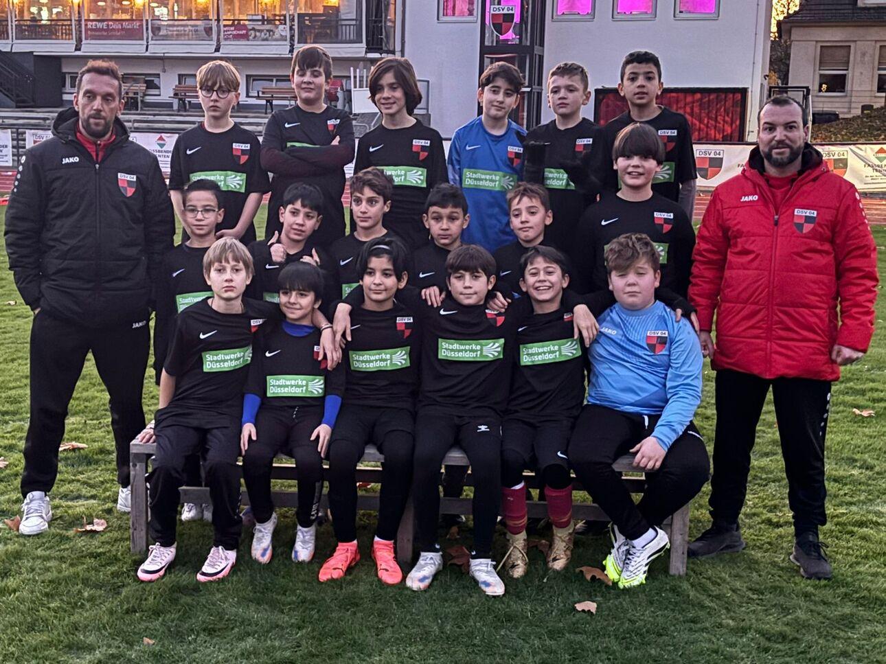 Gruppenshooting von 16 jungen Fu&szlig;ballspielern in schwarz-gr&uuml;nen Trikots, umgeben von zwei Trainern, auf einem Sportplatz.