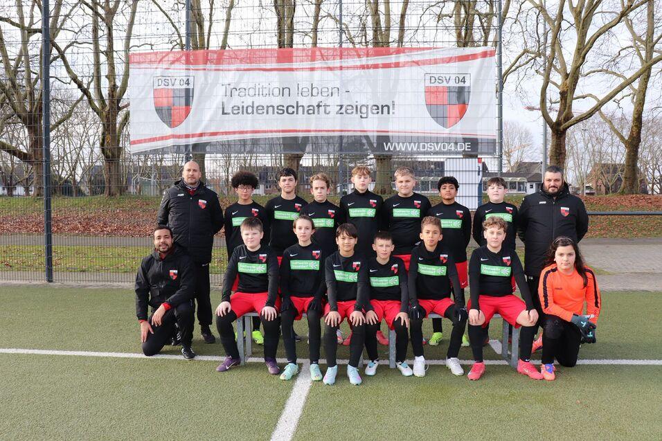 Mannschaftsfoto einer Jugendfußballmannschaft in Schwarz-Rot mit Trainern auf einem Fußballplatz vor einem Banner.