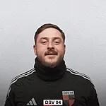 Portr&auml;t eines Mannes in einem Trainingsanzug mit Vereinslogo, l&auml;chelnd vor hellem Hintergrund.