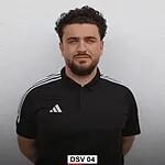 Mann mit lockigem Haar und schwarzem Polohemd steht vor einer wei&szlig;en Wand. Text &bdquo;DSV 04&ldquo; sichtbar.