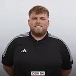 Ein junger Mann mit lockigem Haar tr&auml;gt ein schwarzes T-Shirt mit wei&szlig;en Streifen und l&auml;chelt vor einem neutralen Hintergrund.