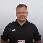 L&auml;chelnder Mann mit kurzen Haaren tr&auml;gt ein schwarzes Adidas-T-Shirt, Hintergrund ist hell und unscharf.