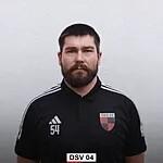 Portr&auml;t eines Mannes mit Bart in schwarzem Shirt mit Logo vor einer grauen Wand.