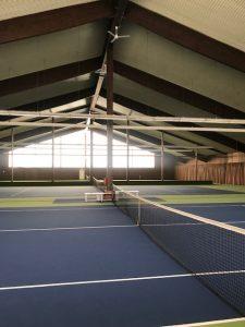 Innenansicht einer Tennishalle mit blauen Pl&auml;tzen, Netz in der Mitte und gro&szlig;en Fenstern im Hintergrund.
