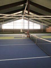 Innenansicht einer Tennishalle mit blauen Pl&auml;tzen, Netz in der Mitte und gro&szlig;en Fenstern im Hintergrund.