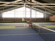 Innenansicht einer Tennishalle mit blauen Pl&auml;tzen, Netz in der Mitte und gro&szlig;en Fenstern im Hintergrund.