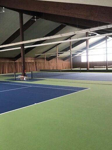 Blick auf mehrere Tennispl&auml;tze in einer Halle mit gr&uuml;nem und blauem Belag sowie gro&szlig;en Fenstern.