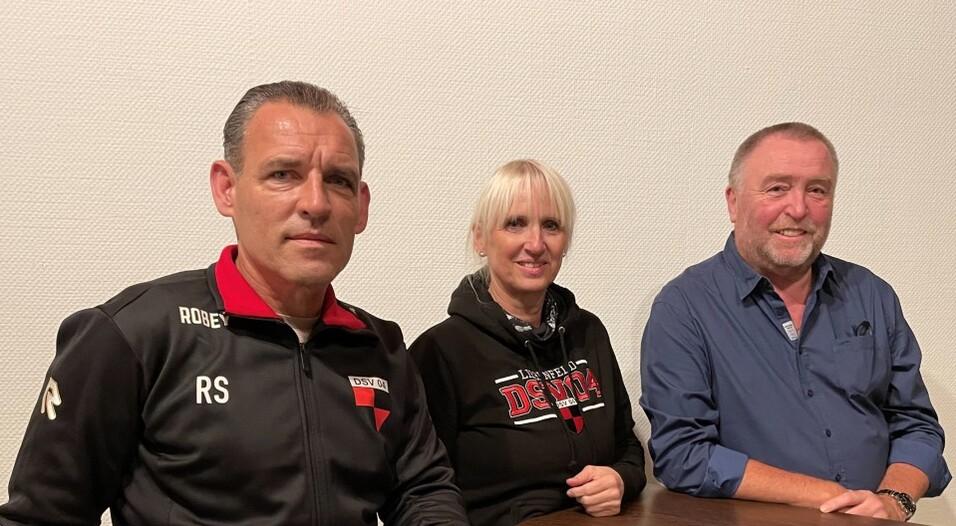 Drei Personen sitzen an einem Tisch, eine Frau in schwarzem Hoodie, ein Mann in Trainingsjacke und ein Mann in blauem Hemd.