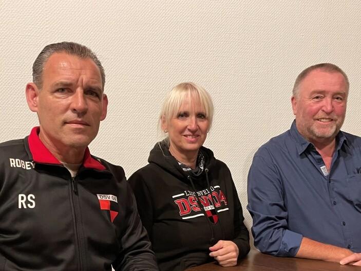 Drei Personen sitzen an einem Tisch, eine Frau in schwarzem Hoodie, ein Mann in Trainingsjacke und ein Mann in blauem Hemd.