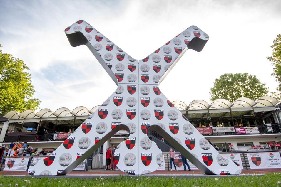 Große, weiße X-Form mit Logos und Emblemen, im Hintergrund ein Stadion und Menschen beim Event.