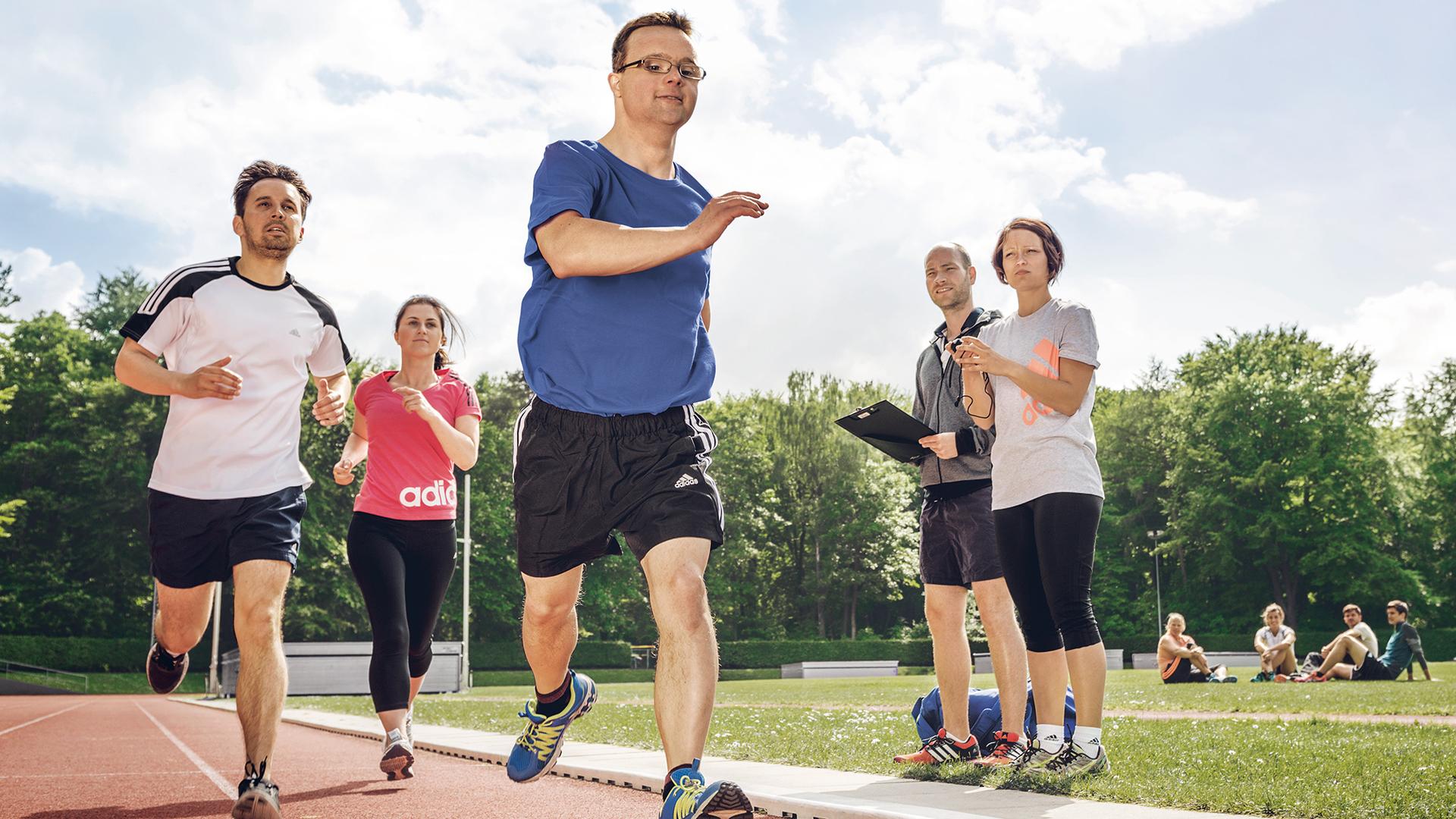 Inklusive Laufgruppe mit Läufer mit Prothese auf der Bahn; zwei Personen beobachten mit Stoppuhr und Klemmbrett.