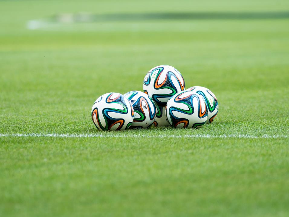 Fünf Fußballbälle liegen auf einem grünen Rasenfeld, sichtbar sind auch der Spielfeldrand und eine verschwommene Hintergrundszene.