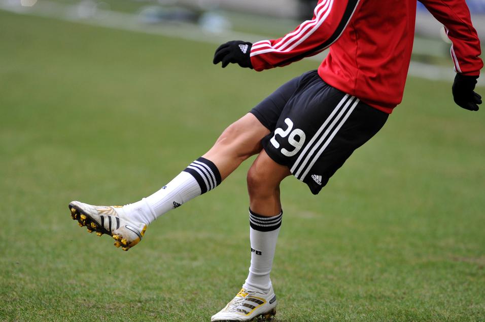 Nahaufnahme eines Fußballspielers mit einem roten Trikot, der auf einem Rasenfeld einen Ball mit dem rechten Fuß spielt.