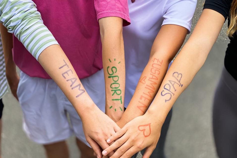 Vier Arme liegen übereinander, mit farbiger Schrift: „TEAM“, „SPORT“, „ZUSAMMEN“, „SPASS“ auf der Haut.