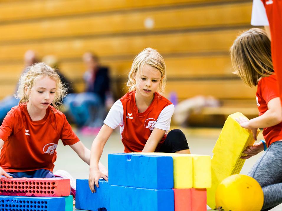 Drei Mädchen im roten Shirt bauen mit bunten Schaumstoffbausteinen in einer Turnhalle.