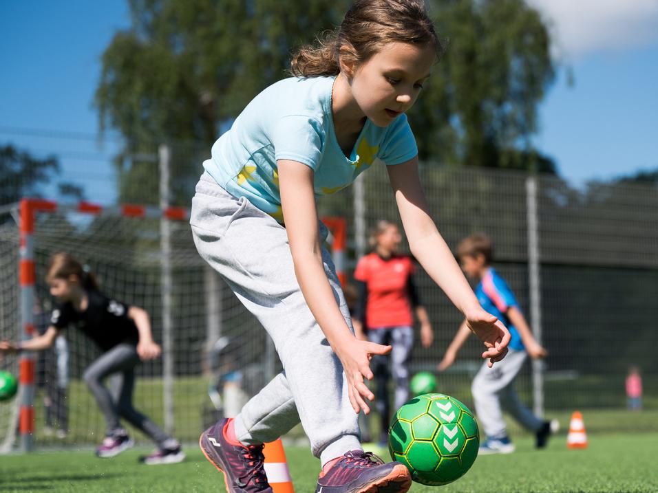 Mädchen in Sportkleidung spielt mit einem grünen Ball auf einem Kunstrasenplatz, andere Kinder trainieren im Hintergrund.