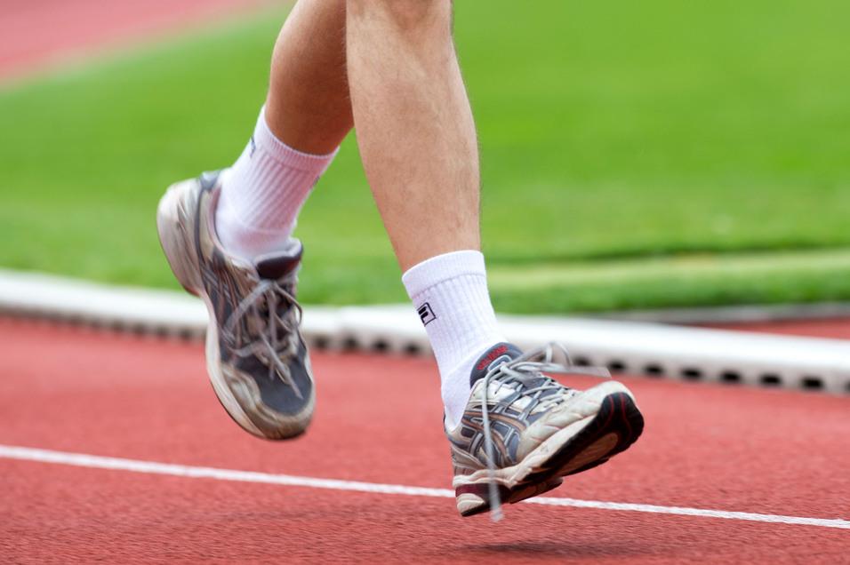 Läufer in grauen Sportschuhen hebt den Fuß auf einer roten Laufbahn über grünem Rasen.