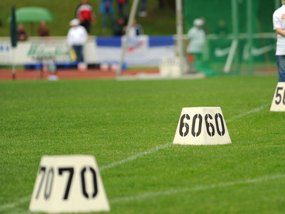 Person mit kurzen Haaren steht auf einem Sportplatz vor nummerierten Markierungen (40, 50, 60, 70) im Gras.