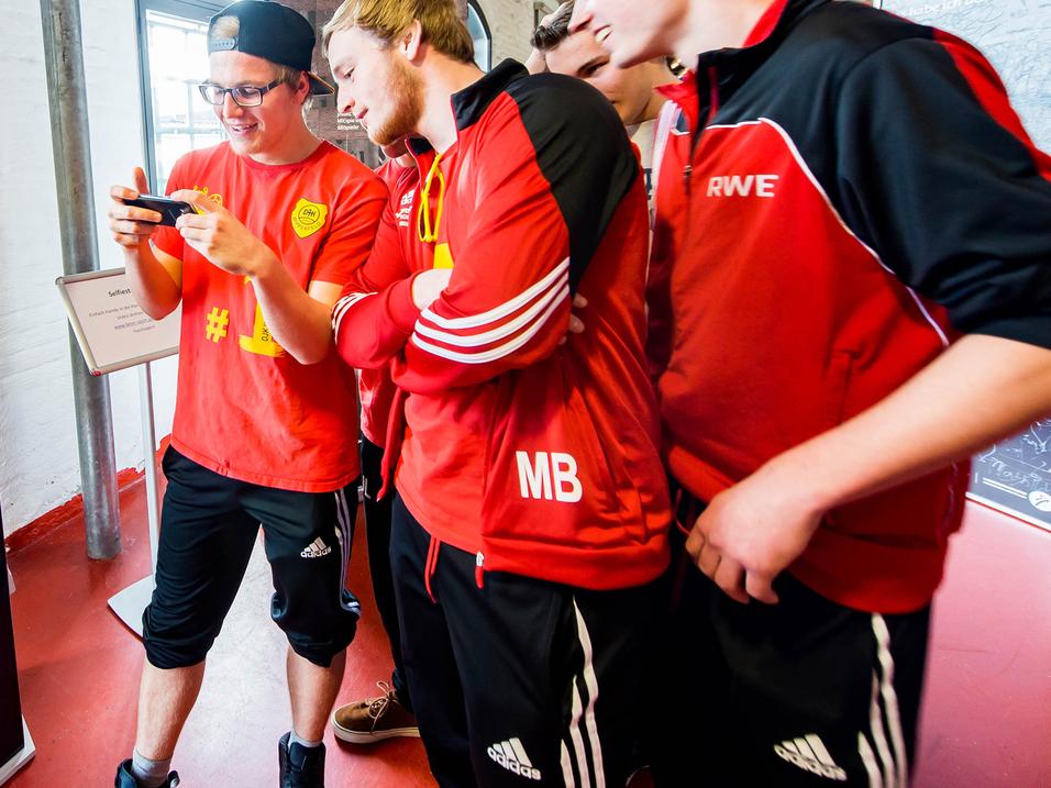 Gruppe junger Sportler in roten Trikots schaut auf ein Smartphone und diskutiert an einem Informationsstand.