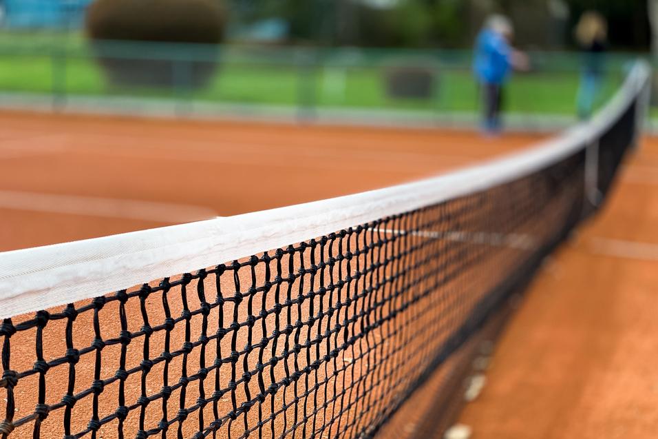 Nahaufnahme eines Tennisnetzes auf einem roten Sandplatz, Hintergrund unscharf mit grünen Bäumen.