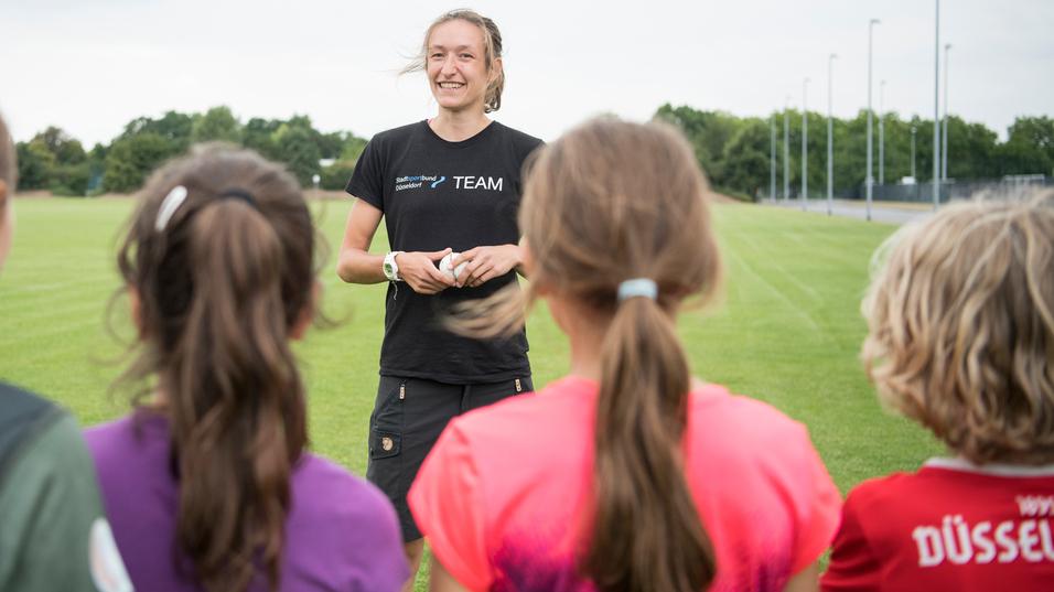 Trainerin spricht mit jungen Sportlerinnen auf einem grünen Fußballfeld unter bewölkten Himmel.