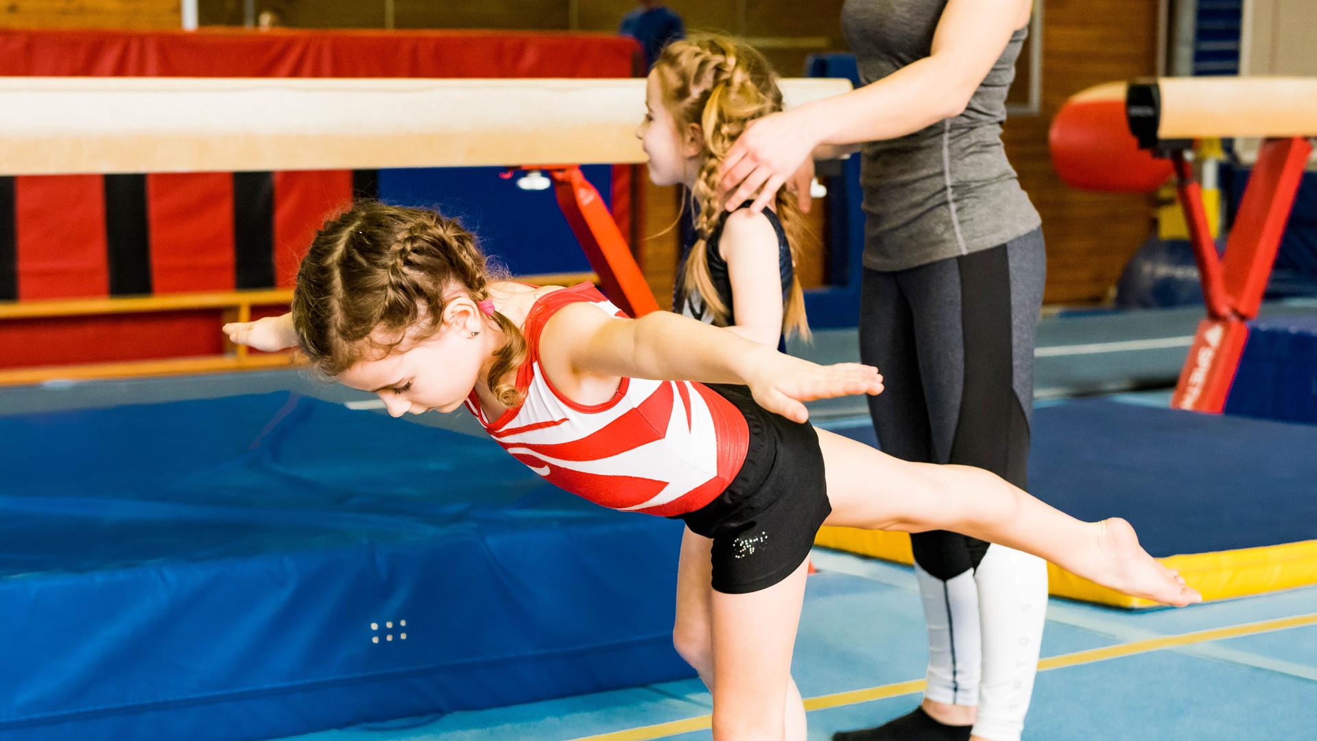 Zwei Mädchen trainieren in einer Turnhalle; eines balanciert auf einem Schwebebalken, während das andere unterstützt.