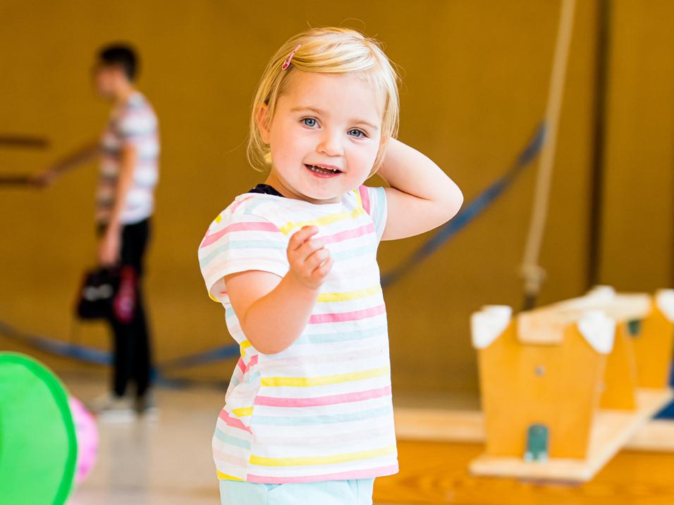 Kleines Mädchen mit blonden Haaren und gestreiftem T-Shirt, das fröhlich in einer Turnhalle spielt.