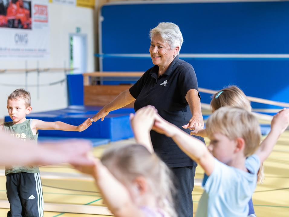 Seniorin leitet Kinder in einer Sporthalle an, während sie ihre Arme in die Höhe strecken.